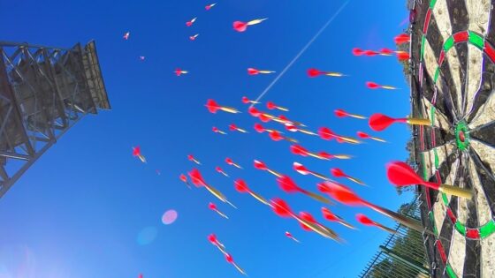 Blue sky with many red-tipped darts flying toward a circular, multicolored target at right; a tall metal tower on the left acts as a backdrop (fairground game).