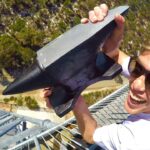 Man wearing sunglasses at the top of a metal tower, smiling, and holding a large black wedge-shaped metal object toward the camera.