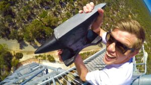 Man wearing sunglasses at the top of a metal tower, smiling, and holding a large black wedge-shaped metal object toward the camera.