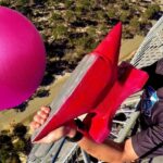 Man wearing sunglasses laughs on a metal platform, holding a red fin and a large pink ball nearby outdoors.