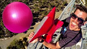 Man wearing sunglasses laughs on a metal platform, holding a red fin and a large pink ball nearby outdoors.