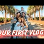 Family of six posing on a sunny palm-lined street with a bold 'OUR FIRST VLOG!' banner across the bottom.