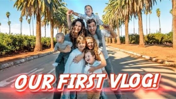 Family of six posing on a sunny palm-lined street with a bold 'OUR FIRST VLOG!' banner across the bottom.