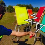 Person in a blue shirt with a neon vest throws a red dart at a row of colorful target boards outdoors. A visible dartboard sits between the boards.