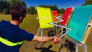 Person in a blue shirt with a neon vest throws a red dart at a row of colorful target boards outdoors. A visible dartboard sits between the boards.