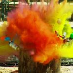 Color powder explosion bursting from spray cans stuck in a tree stump outdoors, creating vivid yellow and orange clouds.