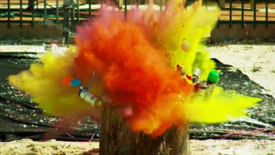 Color powder explosion bursting from spray cans stuck in a tree stump outdoors, creating vivid yellow and orange clouds.