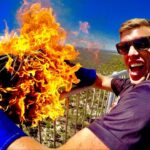 Man on an outdoor observation deck laughs as he holds a burning cloth with large orange flames, wearing gloves and sunglasses.