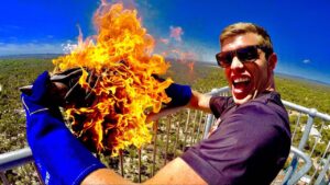 Man on an outdoor observation deck laughs as he holds a burning cloth with large orange flames, wearing gloves and sunglasses.
