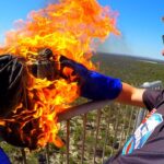 Man wearing sunglasses and blue gloves holds a flame-engulfed bundle on a railing, smiling on a sunny day.