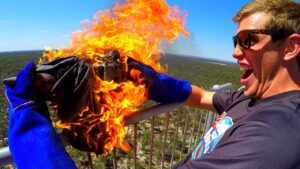 Man wearing sunglasses and blue gloves holds a flame-engulfed bundle on a railing, smiling on a sunny day.