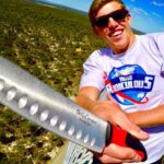 Man wearing sunglasses on a tall outdoor platform, holding a large perforated stainless steel knife toward the camera with a blue sky and landscape in the background.