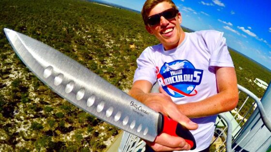 Man wearing sunglasses on a tall outdoor platform, holding a large perforated stainless steel knife toward the camera with a blue sky and landscape in the background.