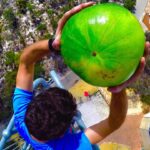 Person in a blue shirt grasps a large bright green watermelon overhead while standing on a tall metal structure, viewed from above to the ground below