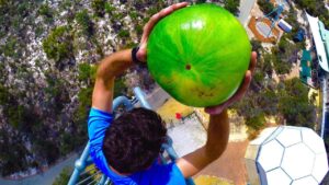 Person in a blue shirt grasps a large bright green watermelon overhead while standing on a tall metal structure, viewed from above to the ground below