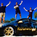 Three men stand on a graffiti-covered black car with arms raised against a bright blue sky, smiling.