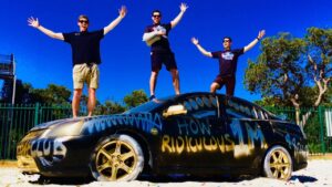Three men stand on a graffiti-covered black car with arms raised against a bright blue sky, smiling.