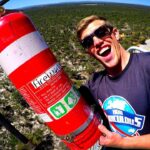 Man wearing sunglasses grins at the camera while holding a large red fire extinguisher outdoors on a viewing platform.