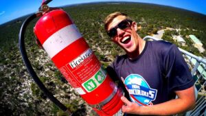 Man wearing sunglasses grins at the camera while holding a large red fire extinguisher outdoors on a viewing platform.