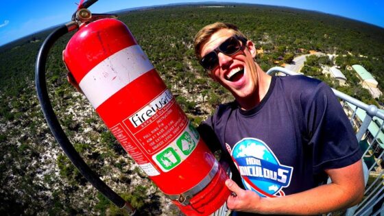 Man wearing sunglasses grins at the camera while holding a large red fire extinguisher outdoors on a viewing platform.
