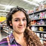 Smiling woman with curly hair in a grocery store aisle, shelves stocked with products behind her.