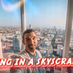 Man selfie in a glass-walled high-rise with a city skyline; red caption 'LIVING IN A SKYSCRAPER' across bottom.
