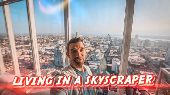 Man selfie in a glass-walled high-rise with a city skyline; red caption 'LIVING IN A SKYSCRAPER' across bottom.