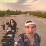 Man in a white cap takes a selfie at the front while a family with a baby stroller walks along a dusty road at sunset.