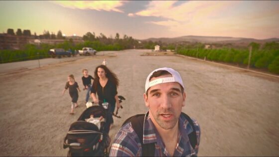 Man in a white cap takes a selfie at the front while a family with a baby stroller walks along a dusty road at sunset.