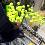Person on a high metal platform dumping a bucket of bright tennis balls into the air, with many balls cascading downward.