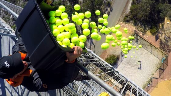 Person on a high metal platform dumping a bucket of bright tennis balls into the air, with many balls cascading downward.