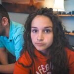 A woman with long wavy hair in an orange hoodie looks at the camera while a man in a turquoise shirt sits beside her, focused on something off-camera in a cluttered room.