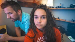 A woman with long wavy hair in an orange hoodie looks at the camera while a man in a turquoise shirt sits beside her, focused on something off-camera in a cluttered room.