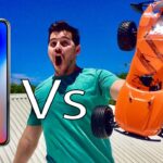Left: a large smartphone screen, right: an orange remote‑controlled car leaping or flipping while a man in a turquoise shirt shouts, with a bold 'Vs' in the center—suggesting a comparison video thumbnail.