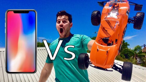 Left: a large smartphone screen, right: an orange remote‑controlled car leaping or flipping while a man in a turquoise shirt shouts, with a bold 'Vs' in the center—suggesting a comparison video thumbnail.