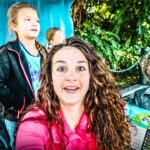 A woman with curly hair takes a selfie with two children outdoors, colorful backdrop and greenery behind them.