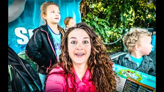 A woman with curly hair takes a selfie with two children outdoors, colorful backdrop and greenery behind them.
