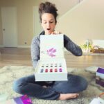 Woman sitting cross-legged on a living room rug opens a white gift box revealing multiple colorful nail polish bottles, surprised expression on her face.
