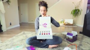 Woman sitting cross-legged on a living room rug opens a white gift box revealing multiple colorful nail polish bottles, surprised expression on her face.