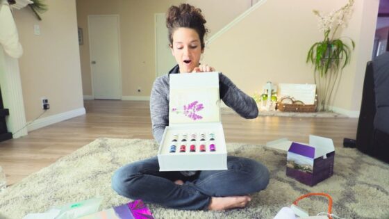 Woman sitting cross-legged on a living room rug opens a white gift box revealing multiple colorful nail polish bottles, surprised expression on her face.
