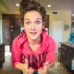 Young woman in a pink hoodie leans toward the camera with a surprised expression, giving two thumbs up in a home kitchen.