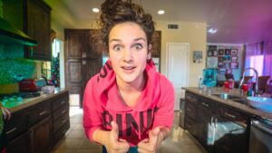 Young woman in a pink hoodie leans toward the camera with a surprised expression, giving two thumbs up in a home kitchen.