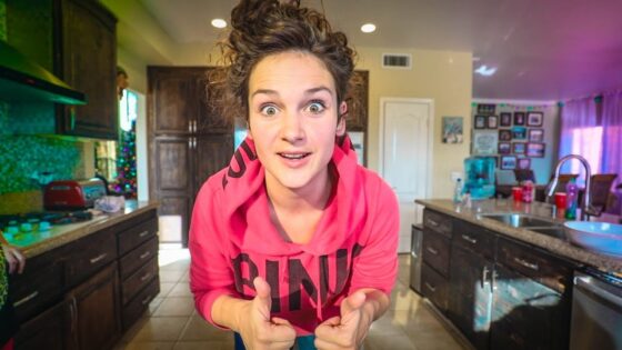Young woman in a pink hoodie leans toward the camera with a surprised expression, giving two thumbs up in a home kitchen.