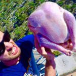 Man in sunglasses selfie-holding a raw turkey high above a landscape, with a road below.