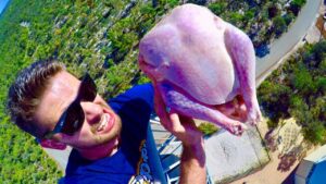 Man in sunglasses selfie-holding a raw turkey high above a landscape, with a road below.