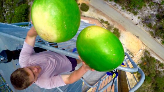 A man in a pink shirt on a tall metal platform, gripping two large green coconuts with a road and landscape far below.