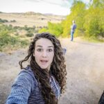 Woman takes a selfie on a dirt road with a white SUV on the right and a man in the distance amid a rural landscape.