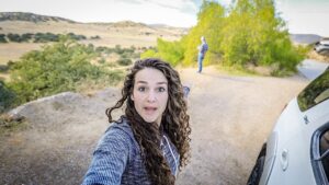 Woman takes a selfie on a dirt road with a white SUV on the right and a man in the distance amid a rural landscape.
