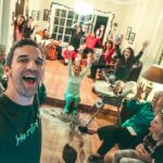 Man in foreground takes a selfie at a festive family Christmas gathering; everyone cheers with raised hands in the living room.