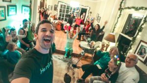Man in foreground takes a selfie at a festive family Christmas gathering; everyone cheers with raised hands in the living room.